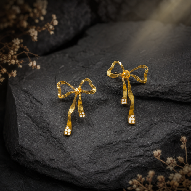 Gold bow-shaped earrings on a dark stone surface with dried flowers.